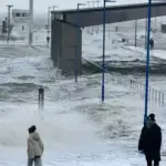 Hohe Sturmflutwellen überspülen die Hafenmole in Dagebüll. Menschen beobachten die aufgewühlte Nordsee bei stürmischem Wetter.
