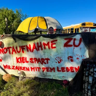 Benthe und Markus Herpich protestieren mit einem Banner an ihrer Containertankstelle in Wyk auf Föhr gegen den geplanten Wegfall der Notaufnahme.