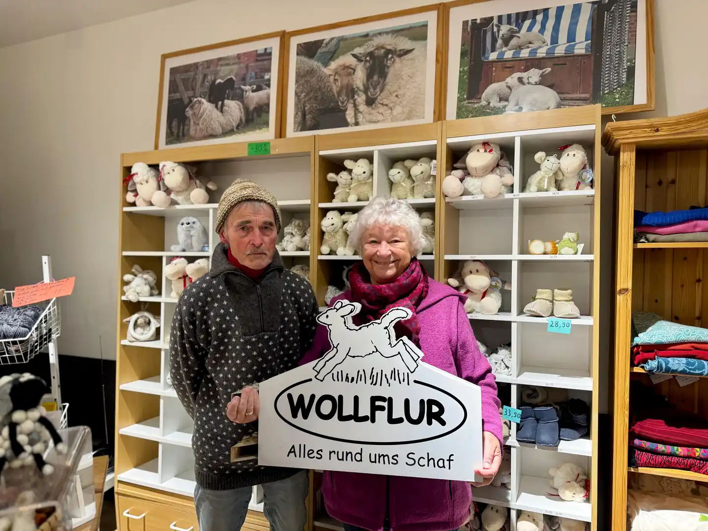 Gaby Brandt und ihr Mann Klaus halten im Wollflur in Wyk auf Föhr das Geschäftsschild mit der Aufschrift „Wollflur – Alles rund ums Schaf“.