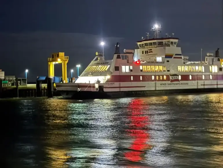 Die beleuchtete W.D.R.-Fähre Uthlande liegt am Samstagabend an der Mole in Dagebüll. Die Lichter spiegeln sich im bewegten Wasser.