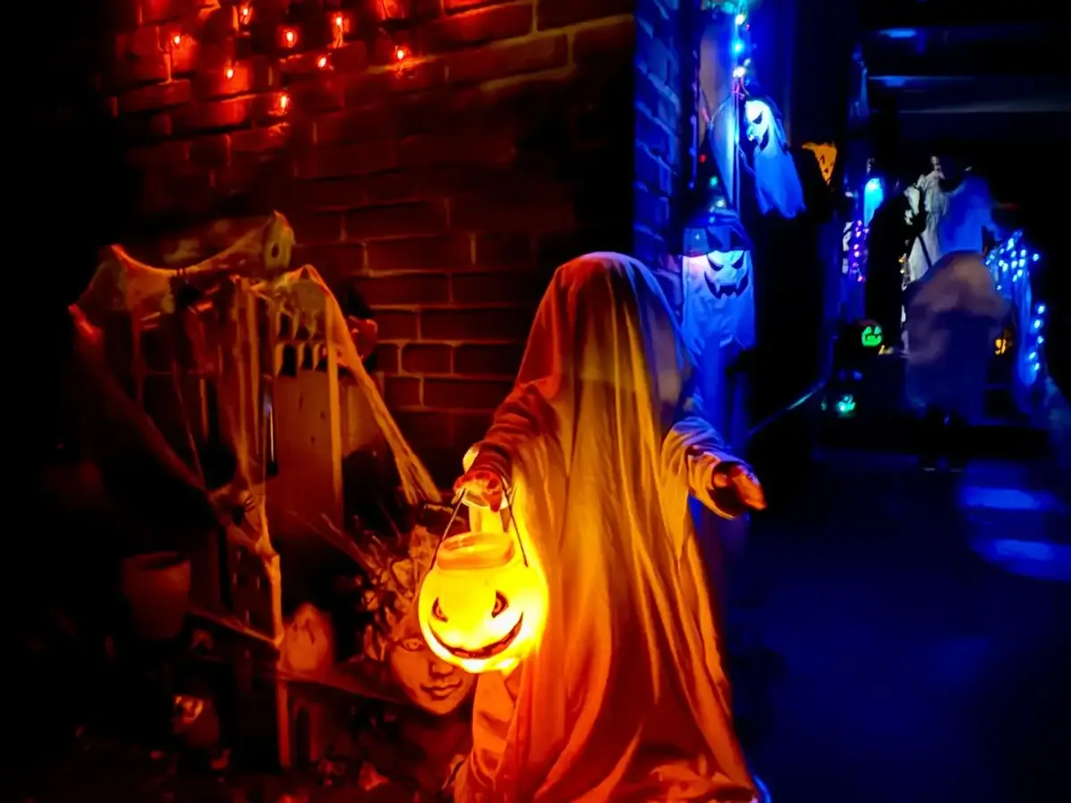 Als Gespenst verkleidetes Kind mit leuchtendem Halloween-Kürbiseimer vor einem gruselig geschmückten Hauseingang.
