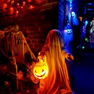 Als Gespenst verkleidetes Kind mit leuchtendem Halloween-Kürbiseimer vor einem gruselig geschmückten Hauseingang.